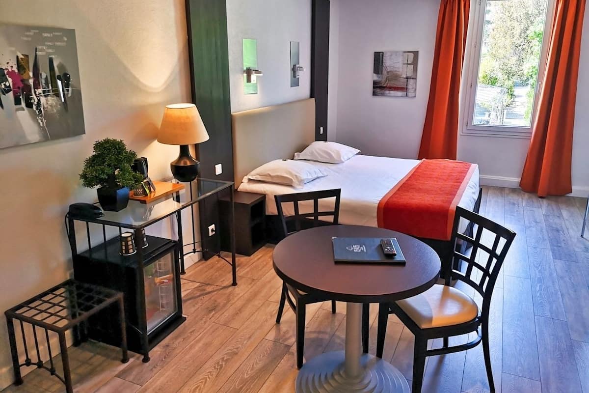 Een hotelkamer met een bed, een ronde tafel met stoelen, een klein bureau met lamp en een raam met gordijnen.