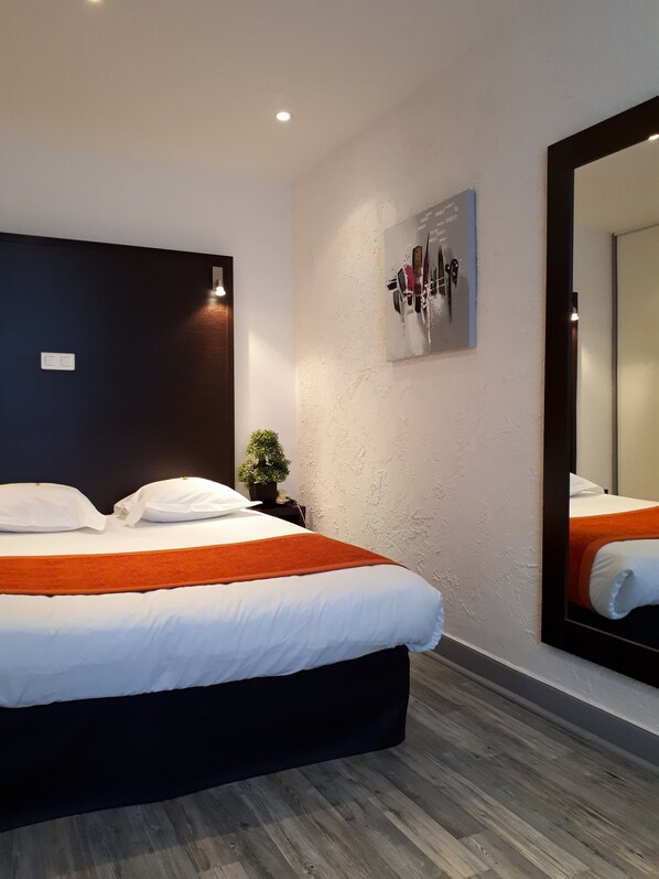 A modern hotel room with a large bed, a dark headboard, a small potted plant, a wall-mounted artwork, and a large mirror.