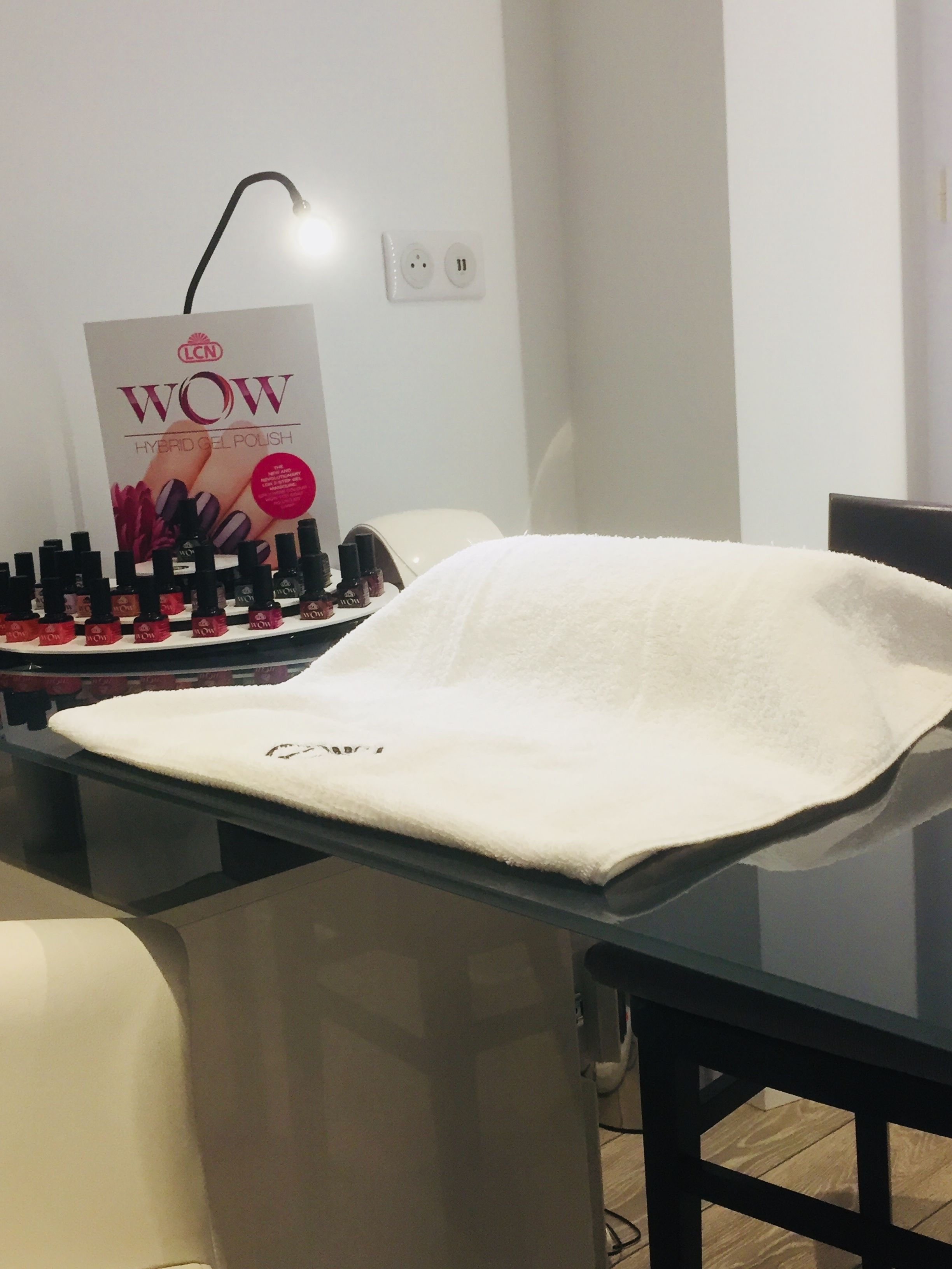 A salon with a white towel on a glass table, a poster for WOW Hybrid Gel Polish, and a lamp.