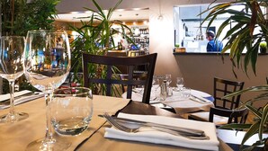 Interior de restaurante com mesas postas para refeições, área de bar ao fundo e plantas em vasos.