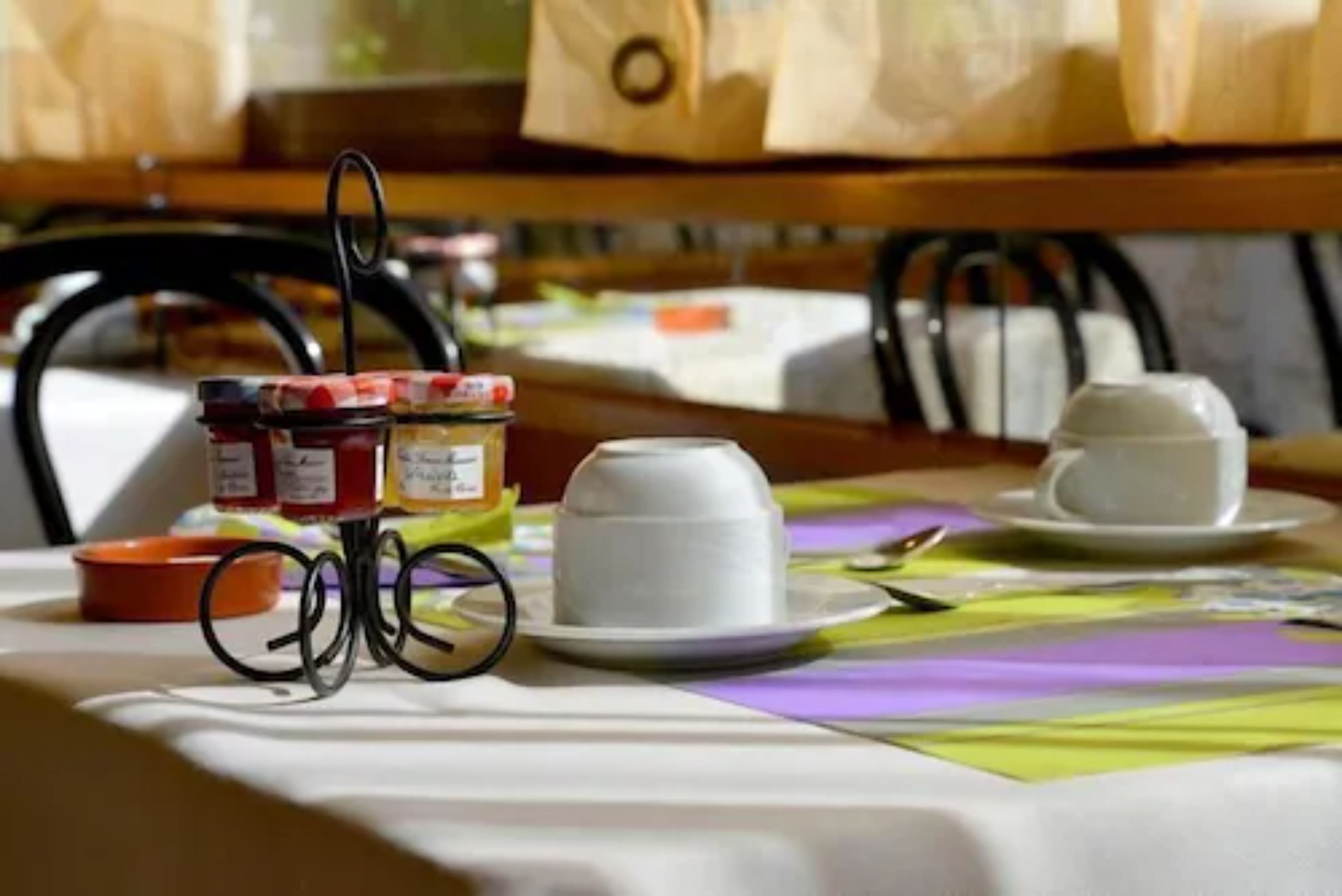 Una mesa con mantel blanco, dos tazas blancas, un frasco de mermelada y un recipiente naranja pequeño.