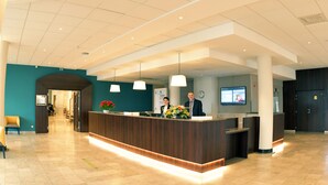 A modern reception area with a wooden reception desk, a digital screen, and a seating area with a chair. - Logis Lyon Est (Saint-Maurice-de-Beynost)
