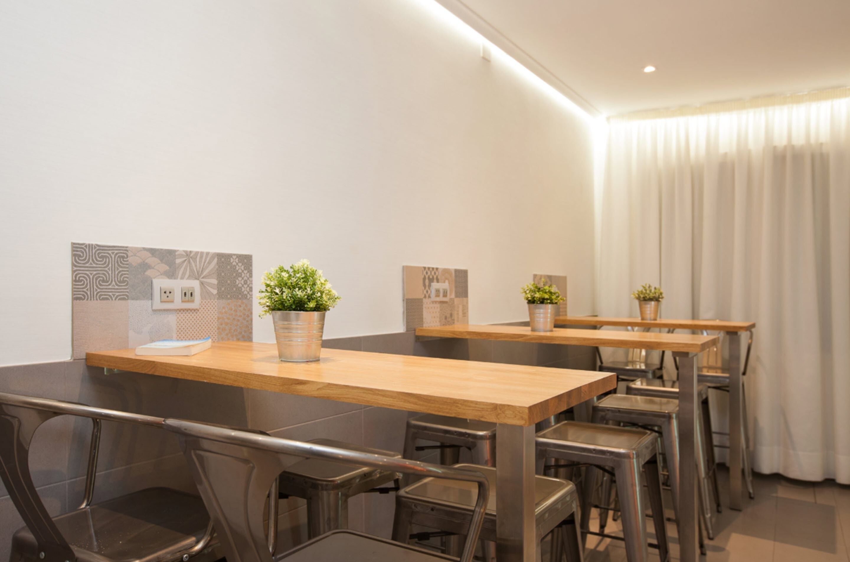 A modern dining area with wooden tables and metal chairs, potted plants, and a white curtain backdrop.
