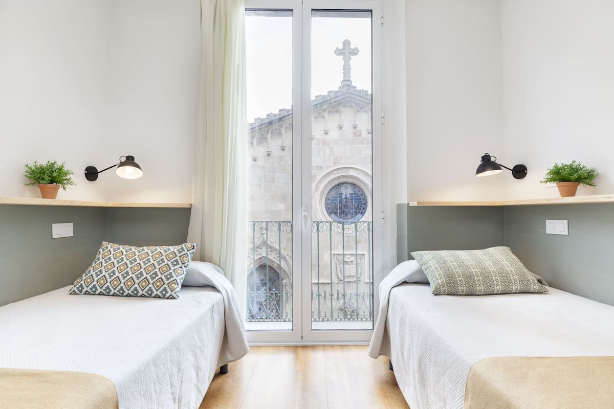 Habitación con dos camas, ventana con vista a un edificio y dos lámparas fijadas a la pared.