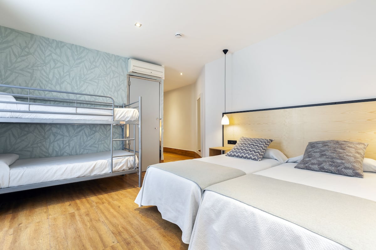 Una habitación con una cama litera, una cama individual y piso de madera. Se observa una puerta, una mesita de noche y una lámpara.