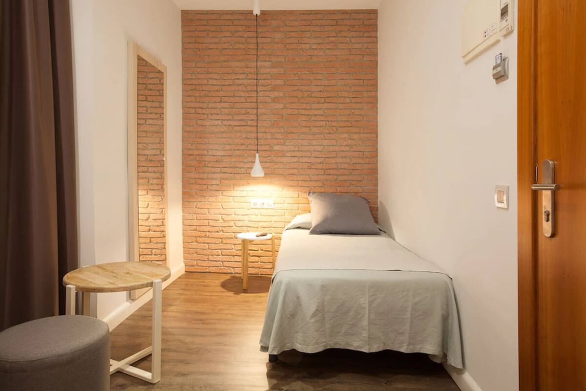Un dormitorio pequeño y acogedor con pared de ladrillo, una cama individual, una mesita de noche, una lámpara colgante y una puerta de madera.