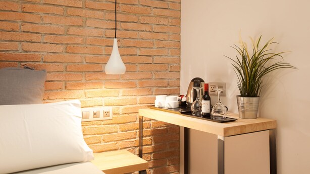 Une chambre moderne avec un mur en briques, un luminaire suspendu, une petite table en bois sur laquelle est posée une plante en pot, et un lit avec des oreillers.