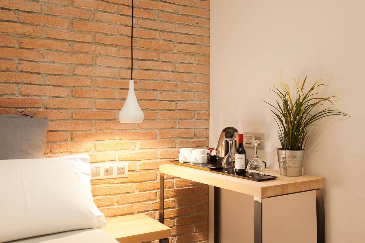 Una habitación moderna con pared de ladrillo, lámpara colgante, una pequeña mesa de madera con planta en maceta y una cama con almohadas.