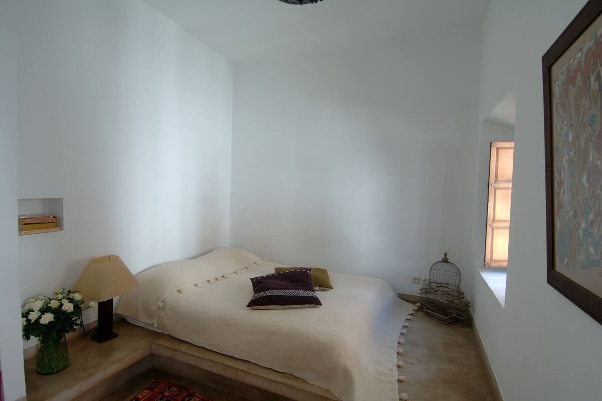 Een compacte, keurig opgemaakte slaapkamer met een bed, een lamp, een vaas met bloemen en een vogelkooi.