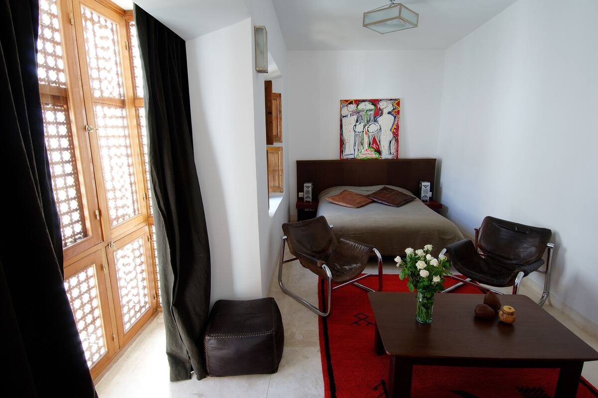 Een hotelkamer met een bed, twee stoelen, een tafel met een bloevas en een rood tapijt.