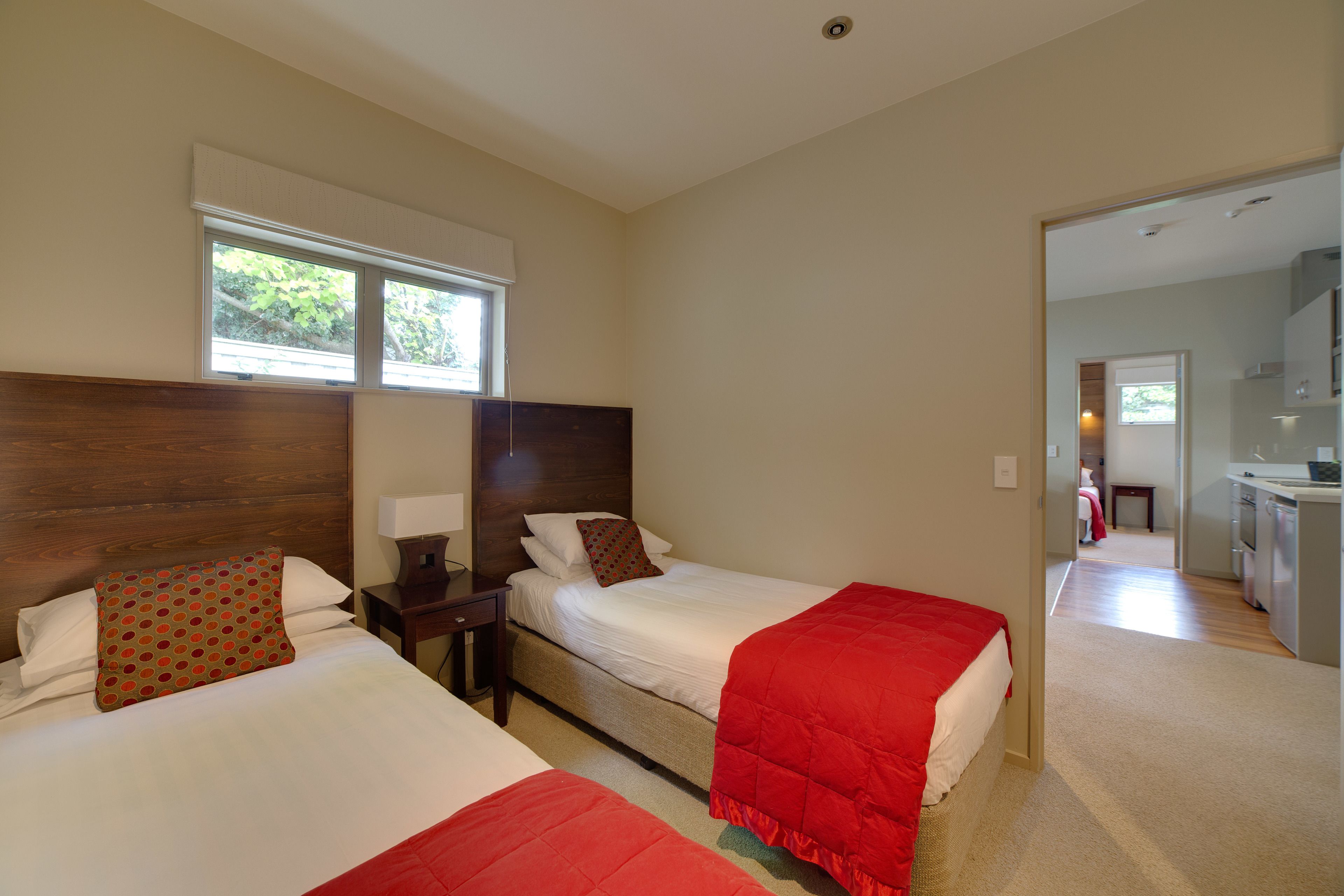 A hotel room with two beds, a wooden headboard, a window with blinds, a nightstand, and a view into the adjacent kitchen area.