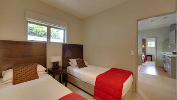 A hotel room with two beds, a wooden headboard, a window with blinds, a nightstand, and a view into the adjacent kitchen area.