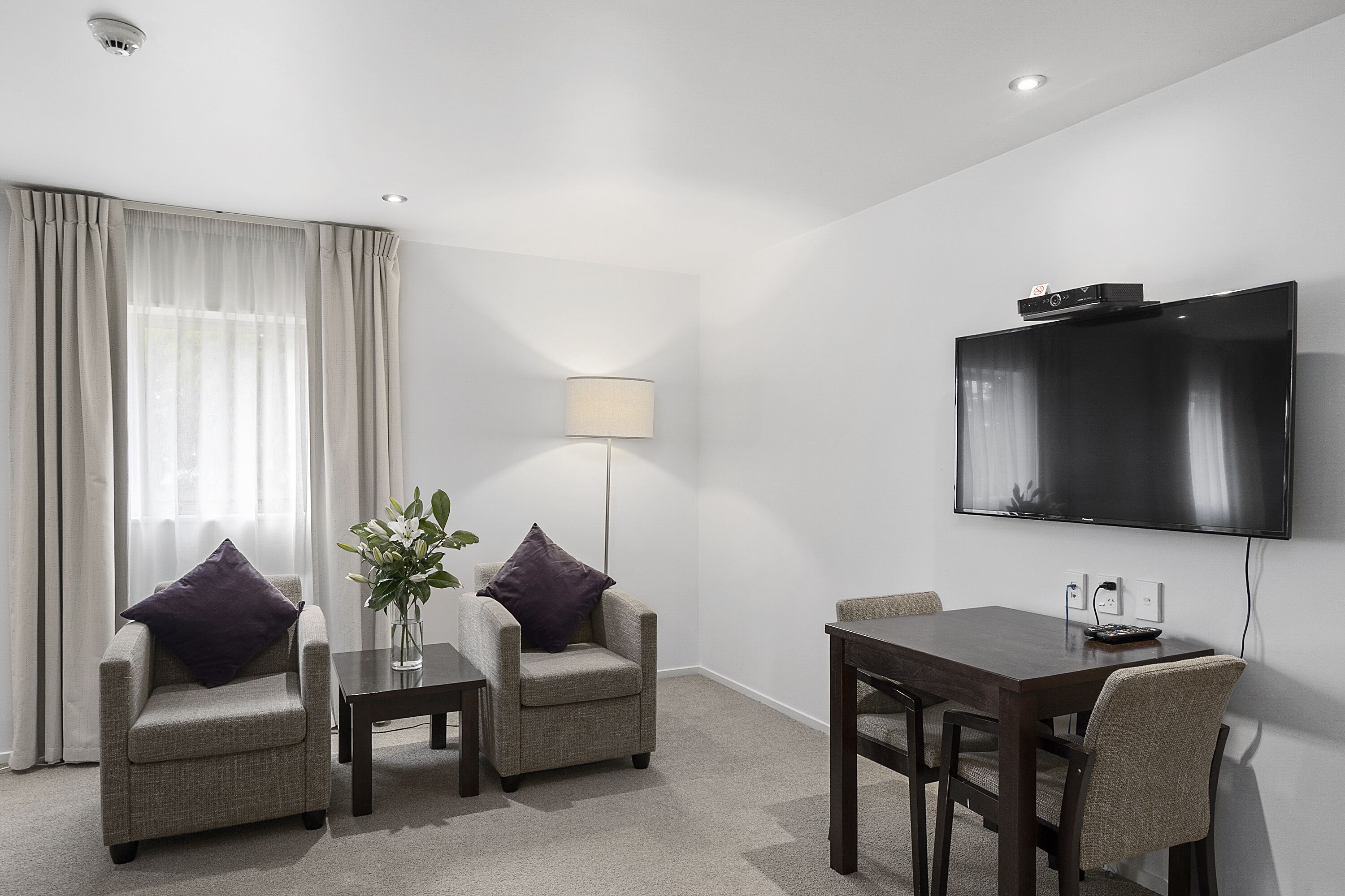 A hotel room with a flat-screen TV mounted on the wall, two armchairs with cushions, a small table, a lamp, and a dining table with chairs.