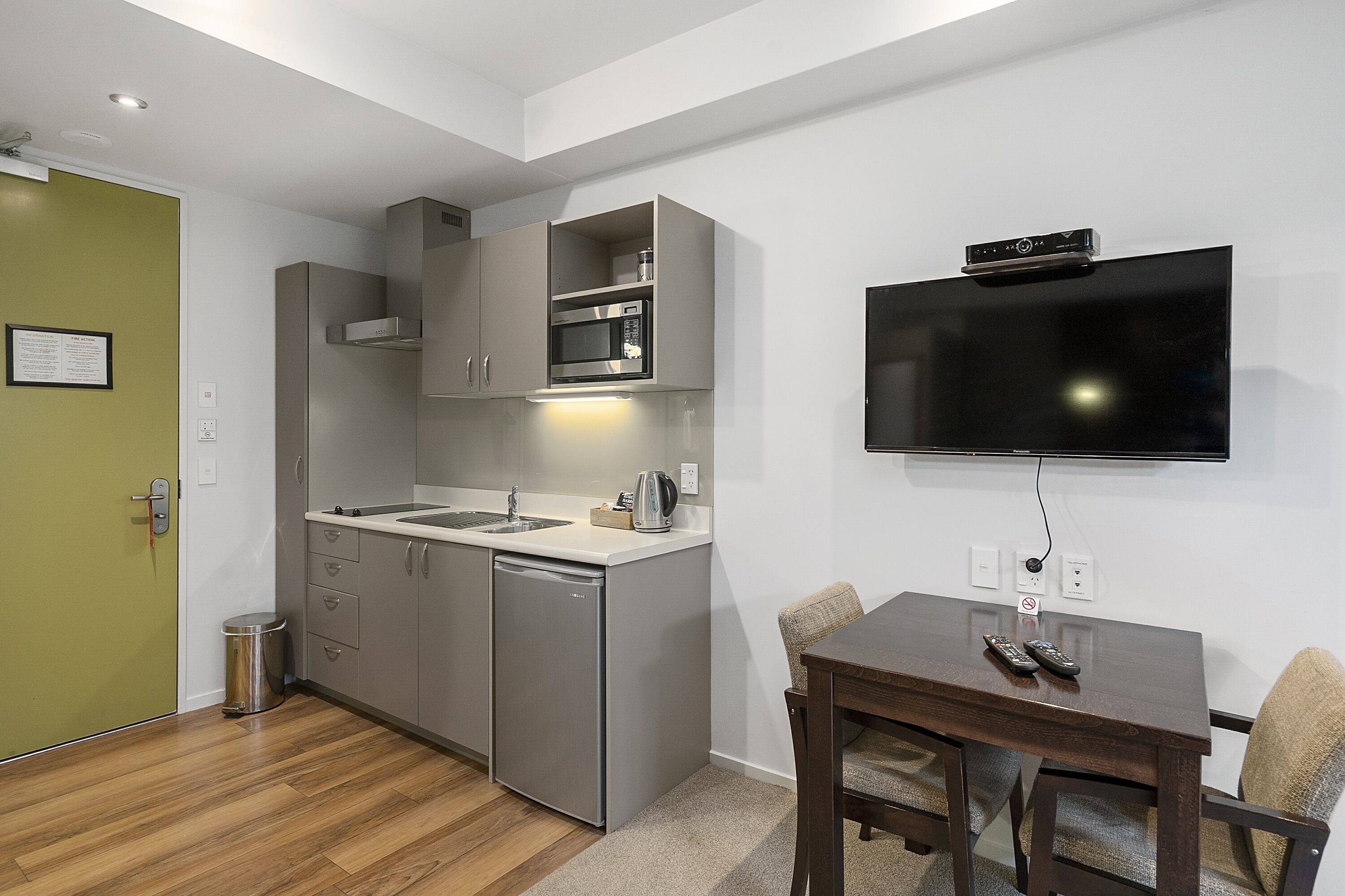 Eine kleine Küche mit Mikrowelle, Spüle und kleinem Kühlschrank. Ein Esstisch mit Stühlen steht neben einem an der Wand montierten Fernseher.