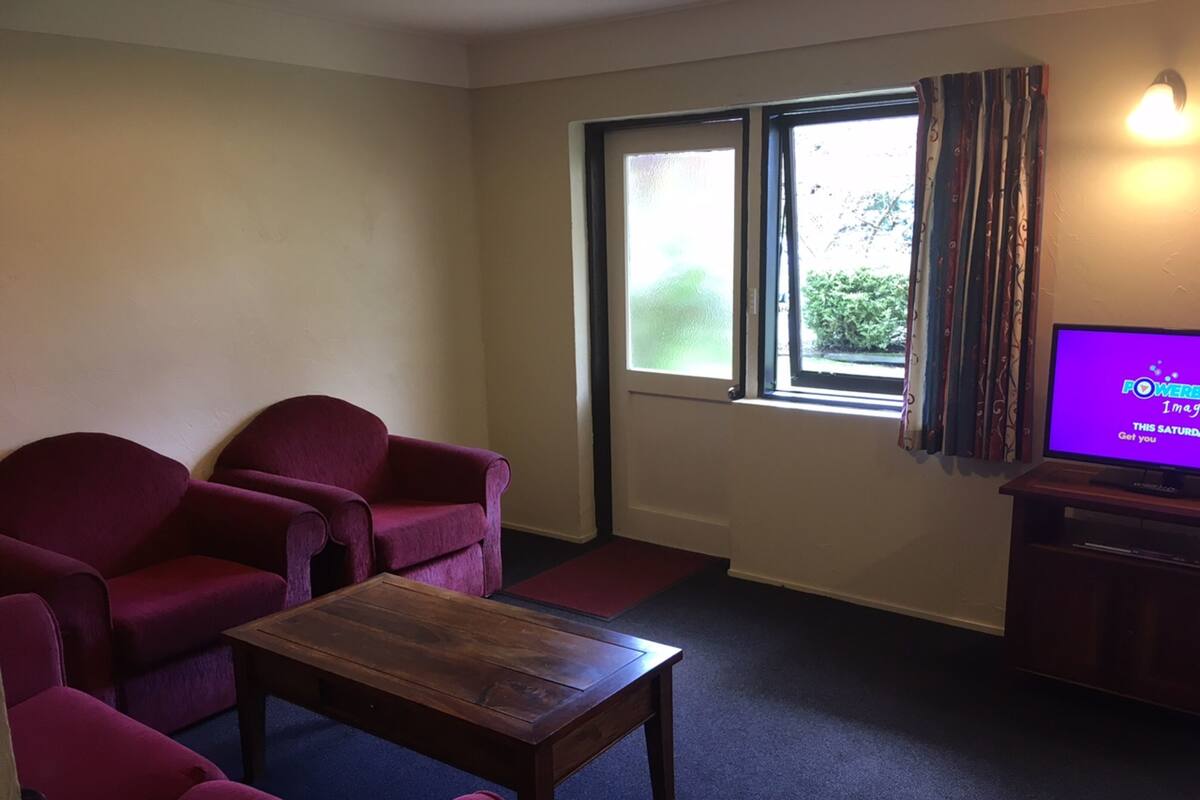 A living room with two red armchairs, a wooden coffee table, a television on a stand, and a window with curtains.