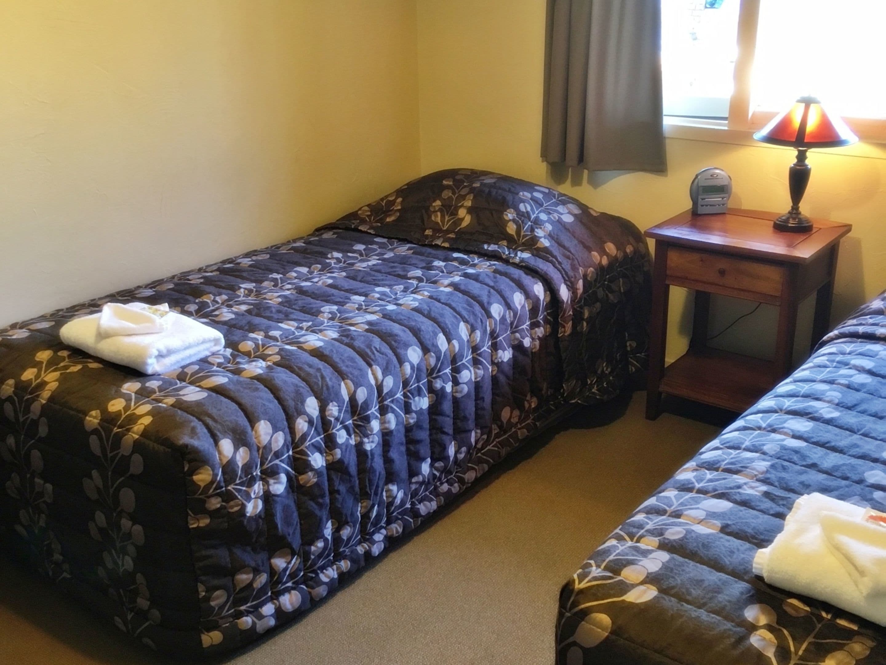 A room with two beds, each with a floral-patterned duvet and a folded towel on the bed. There is a nightstand with a lamp and a clock on it.