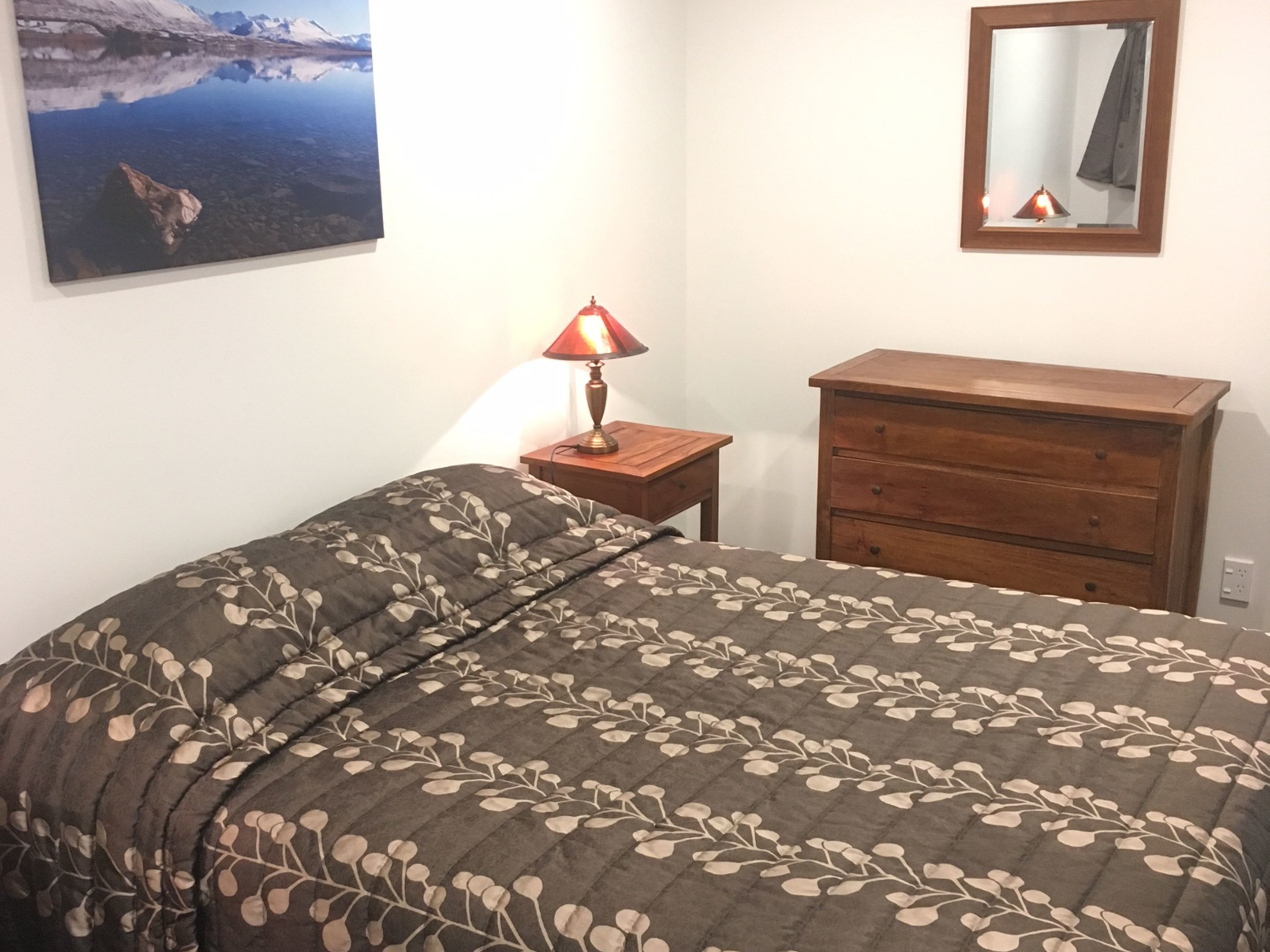 A bedroom with a bed, a dresser, a nightstand with a lamp, a wall-mounted mirror, and a painting of a landscape.