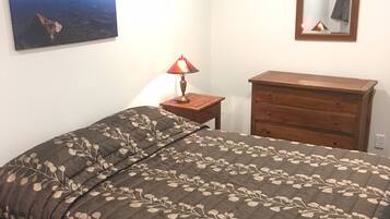 A bedroom with a bed, a dresser, a nightstand with a lamp, a wall-mounted mirror, and a painting of a landscape.