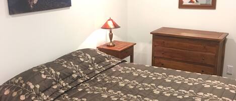 A bedroom with a bed, a dresser, a nightstand with a lamp, a wall-mounted mirror, and a painting of a landscape.