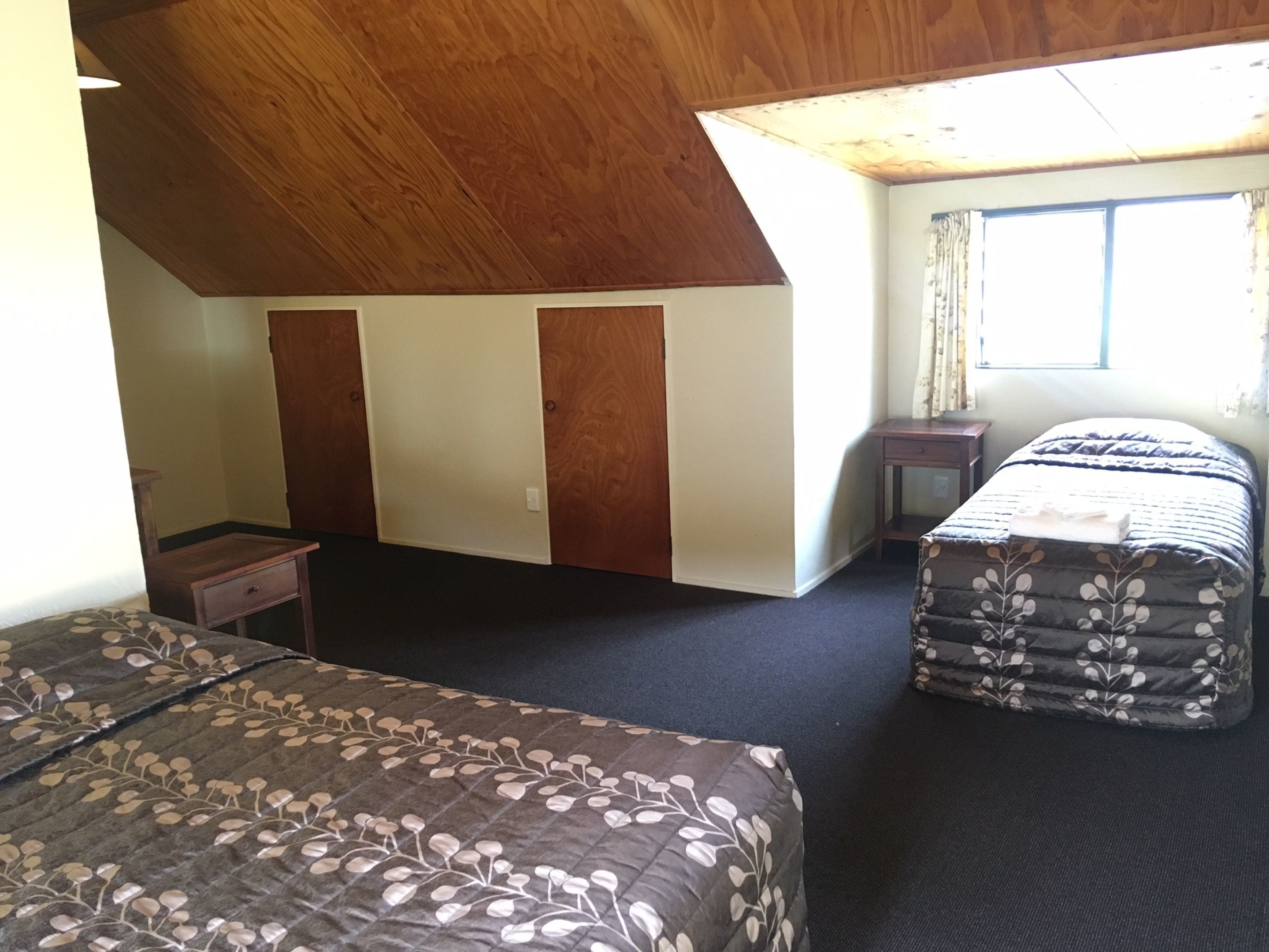 A bedroom with two beds, a wooden ceiling, a window with curtains, and a small bedside table.