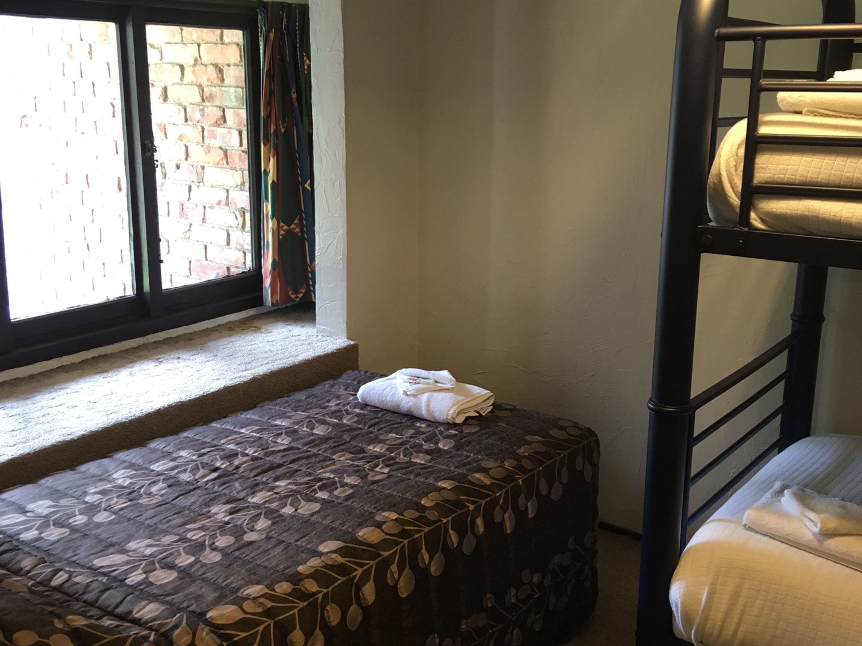 Une petite pièce, de taille modeste, comprenant un lit simple, une fenêtre donnant sur un mur de briques et un lit superposé adossé au mur.