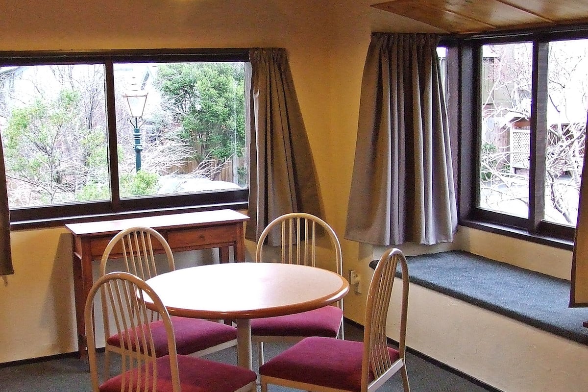 A room with a round table and four chairs, large windows with curtains, and a view of trees and a utility pole outside.
