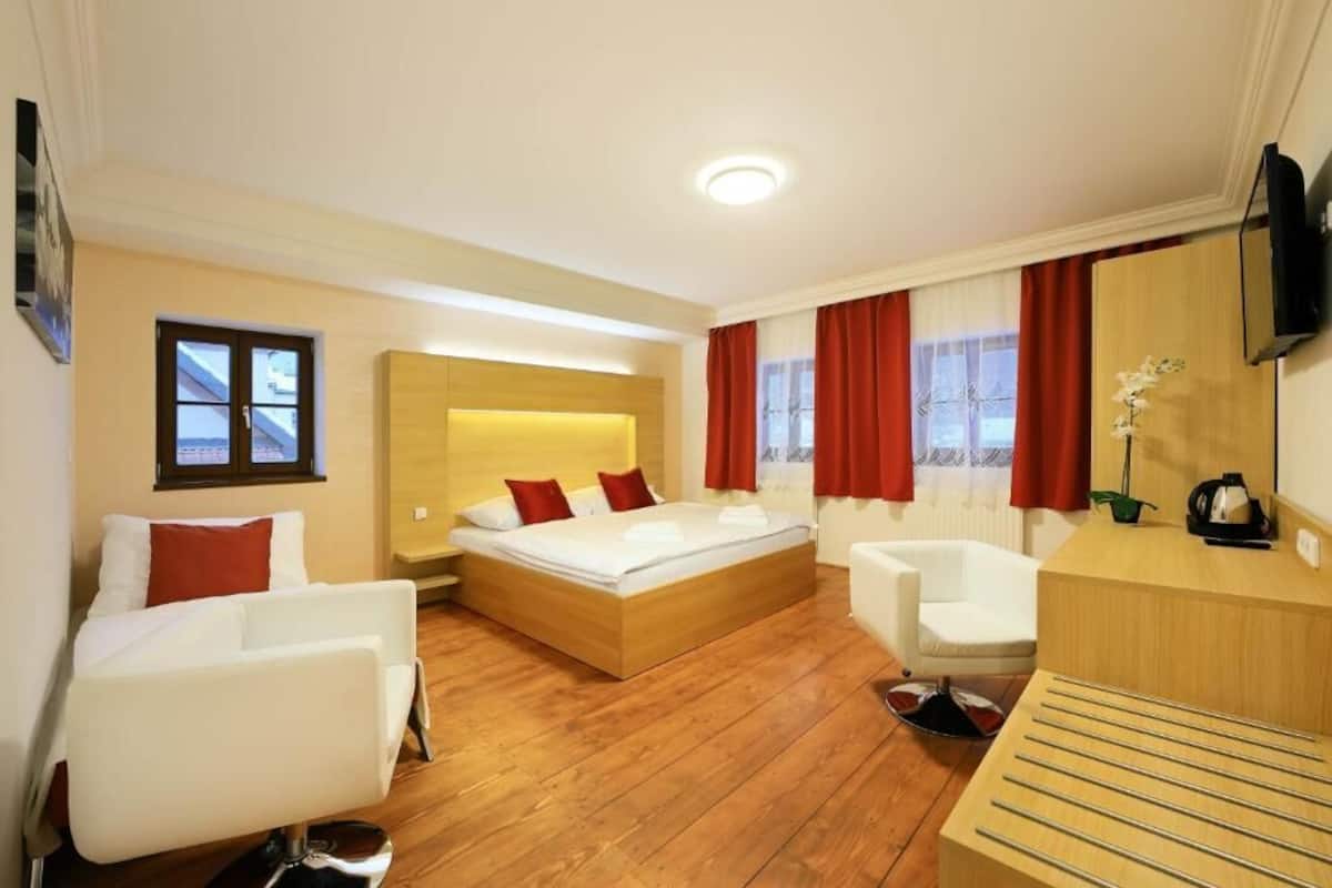 Una habitación de hotel moderna con suelo de madera, una cama con almohadas rojas, un sillón blanco, un escritorio con un hervidor y un televisor montado en la pared.
