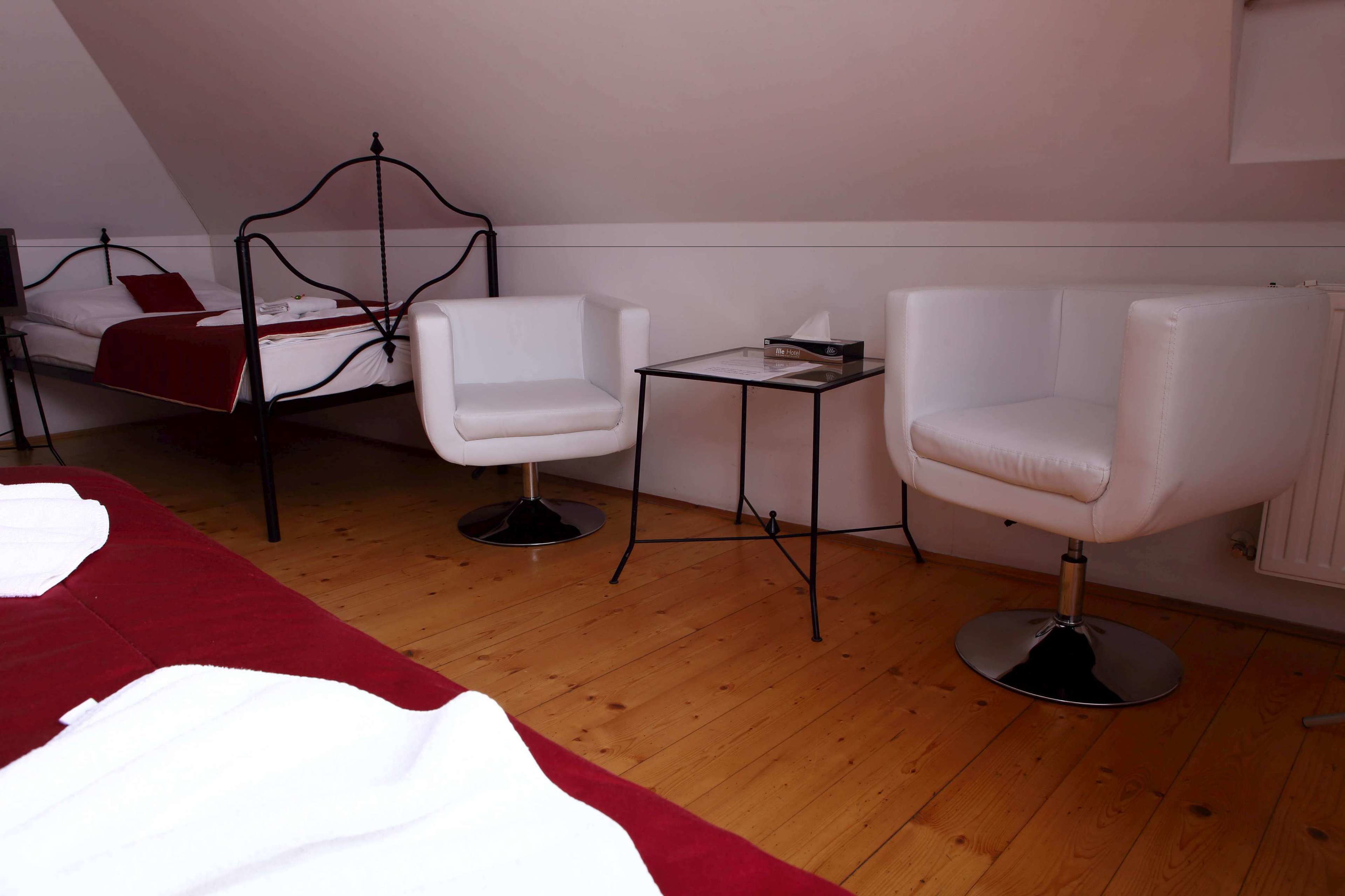 Un dormitorio con una cama, dos sillas blancas, una mesita y una colcha roja.