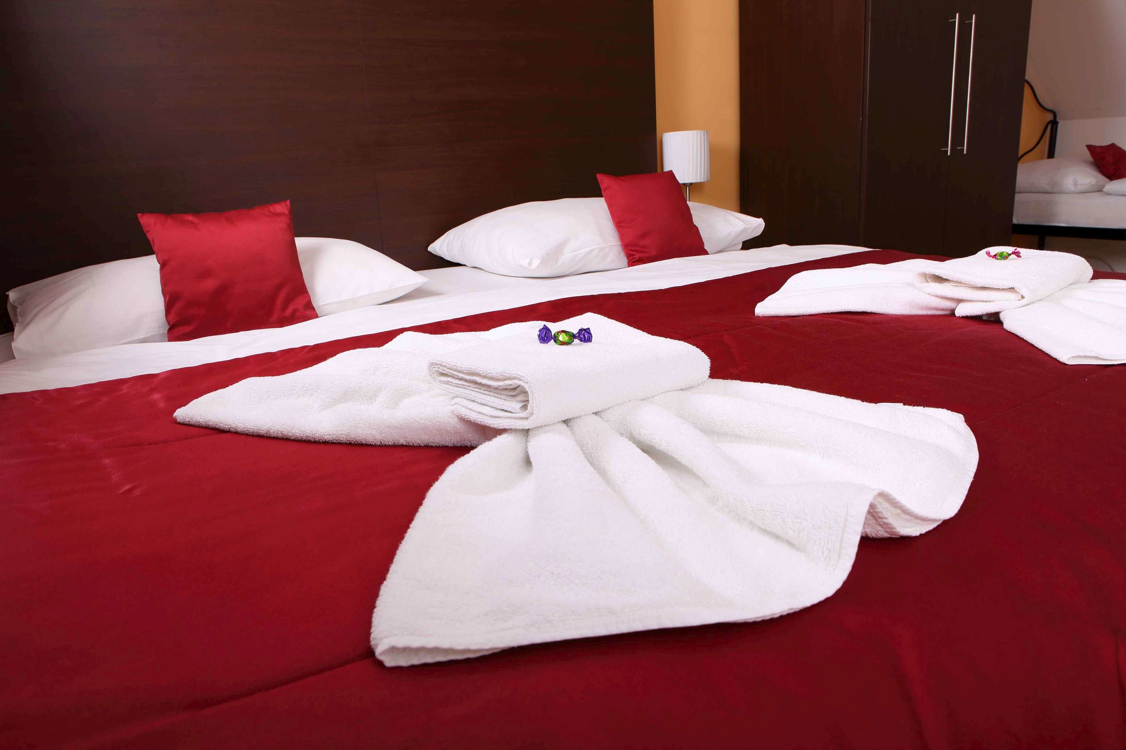 Ett hotellrum med en stor säng, röda sängkläder, vita kuddar och prydligt hopvikta handdukar på sängen.