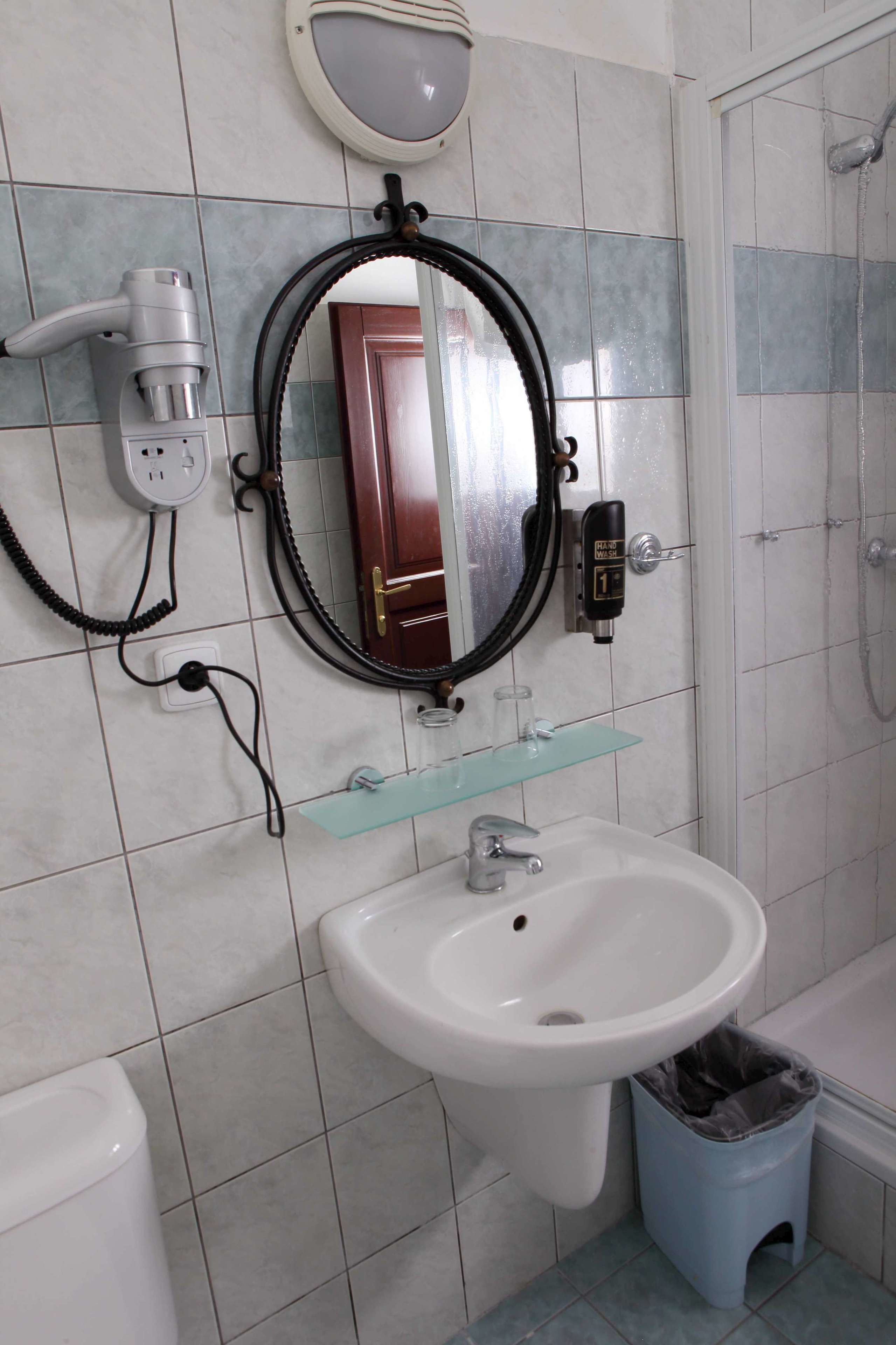 Un baño con lavamanos blanco, espejo, secador de pelo y cubo de basura.