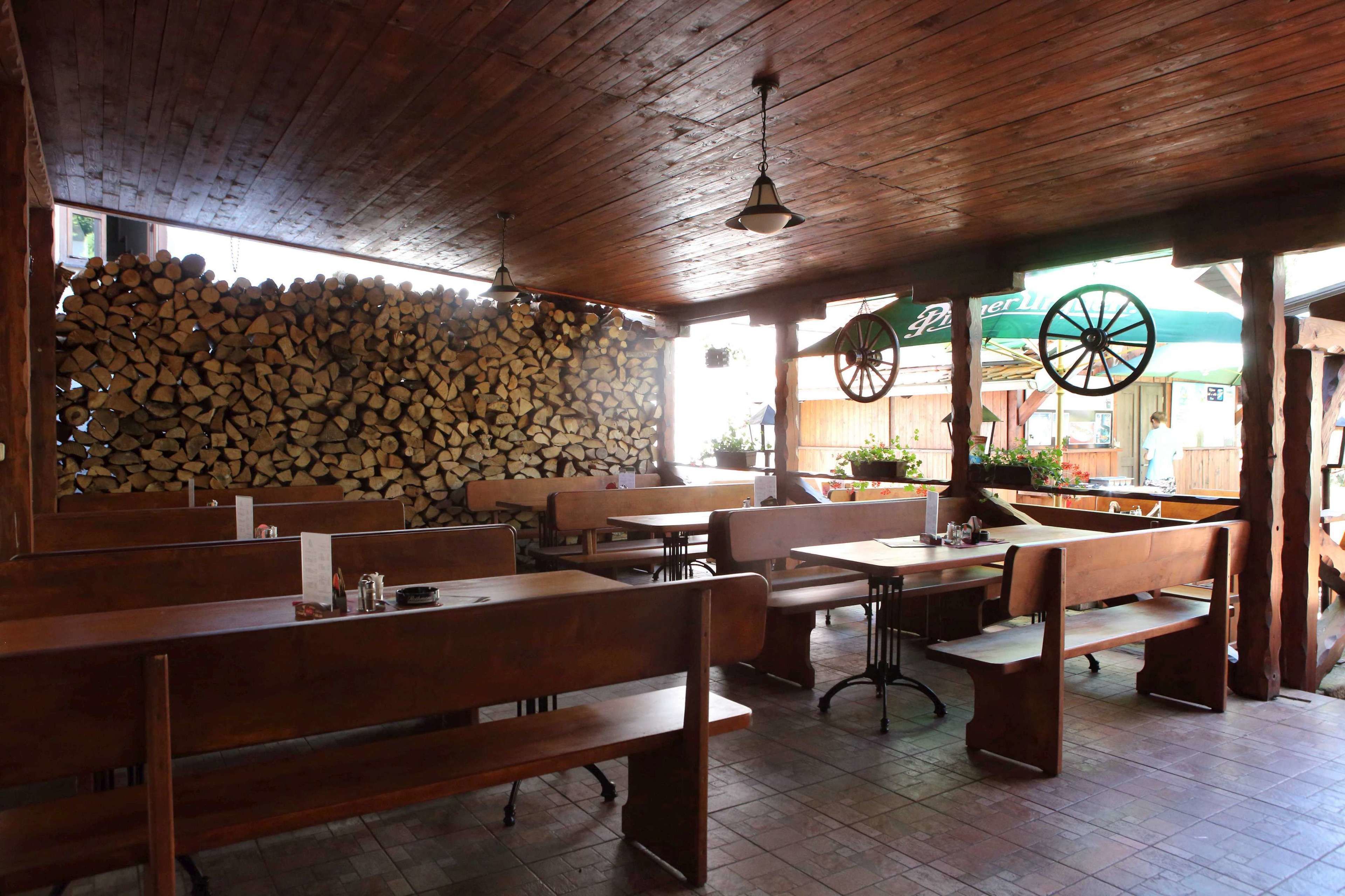 Un interior rústico de madera con bancos y mesas, una pared de piedra con leña apilada y una rueda colgando del techo.