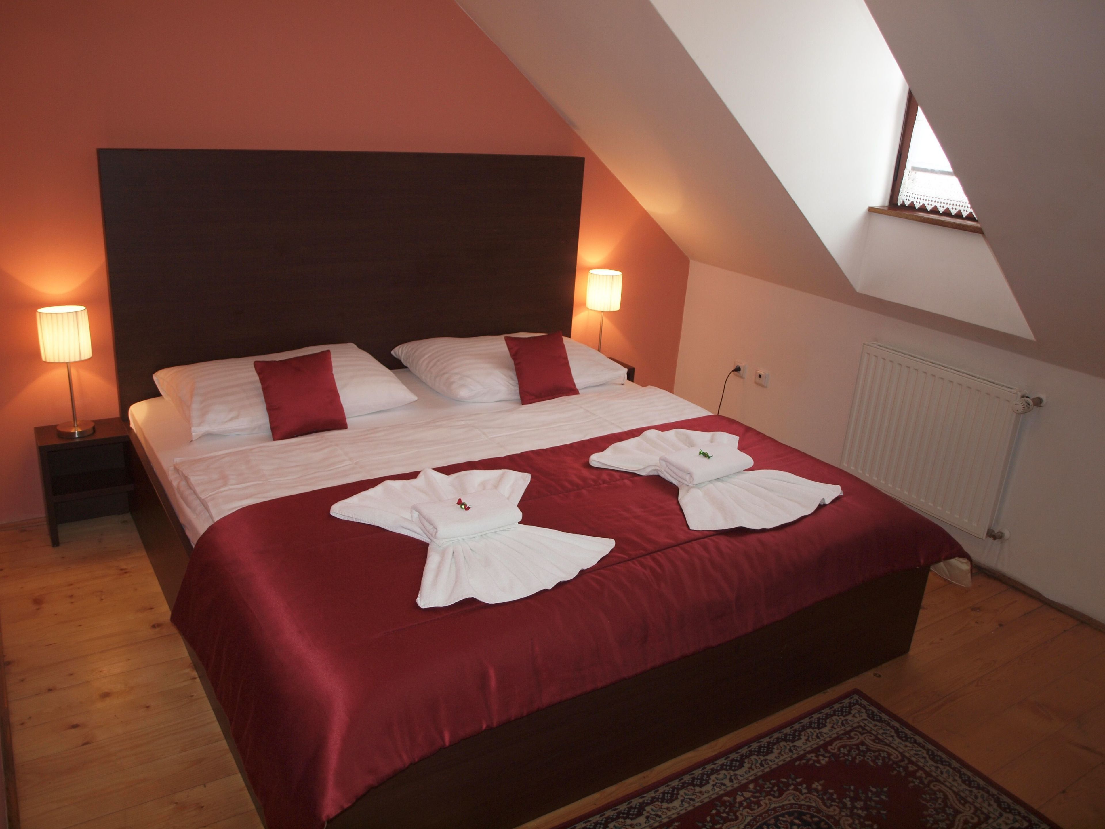 Una cama doble con colcha roja y almohadas blancas, dos lámparas de noche y una ventana con vista al cielo.