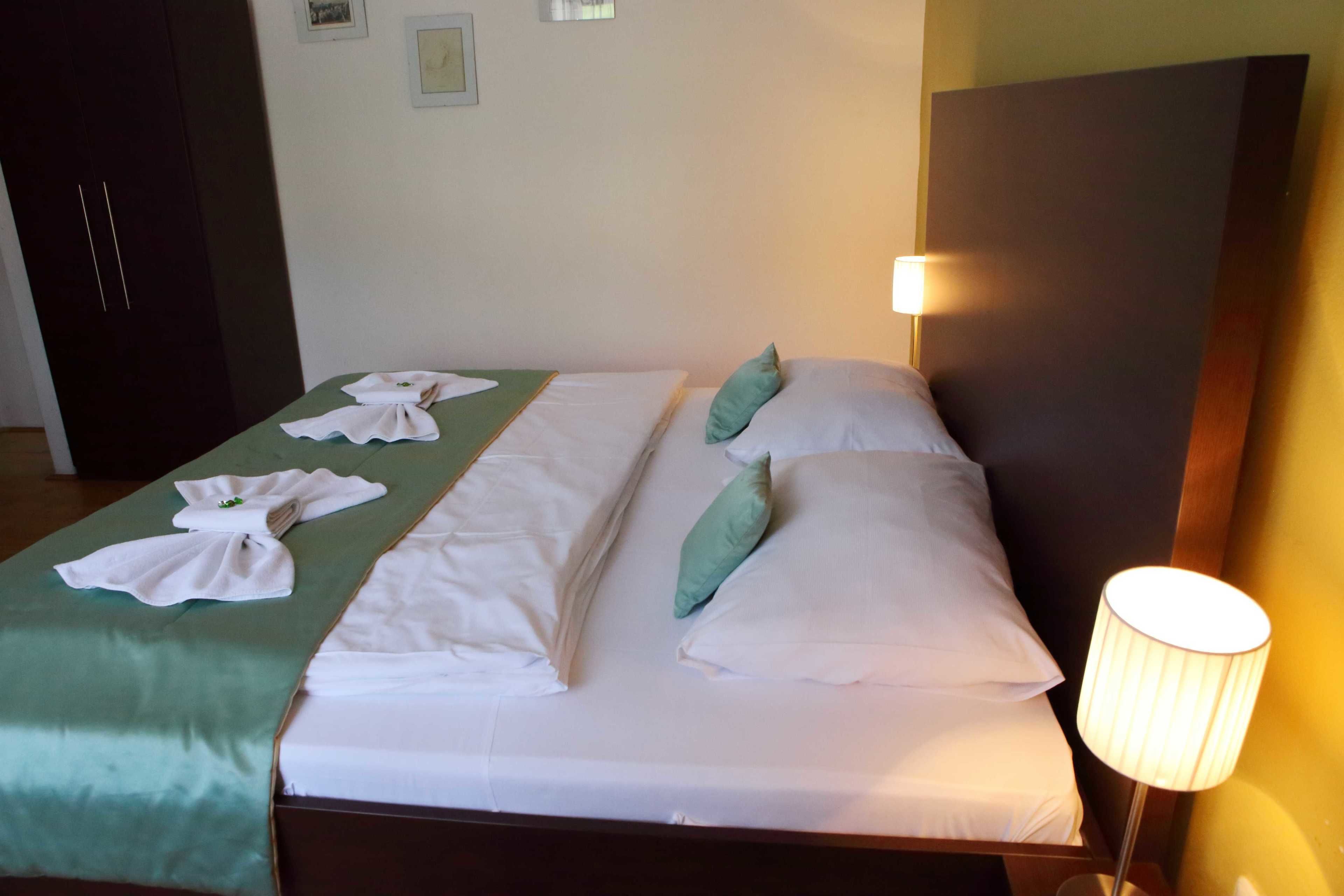 Una cama doble con sábanas blancas, una colcha verde y dos almohadas. Hay una lámpara al lado derecho de la cama.