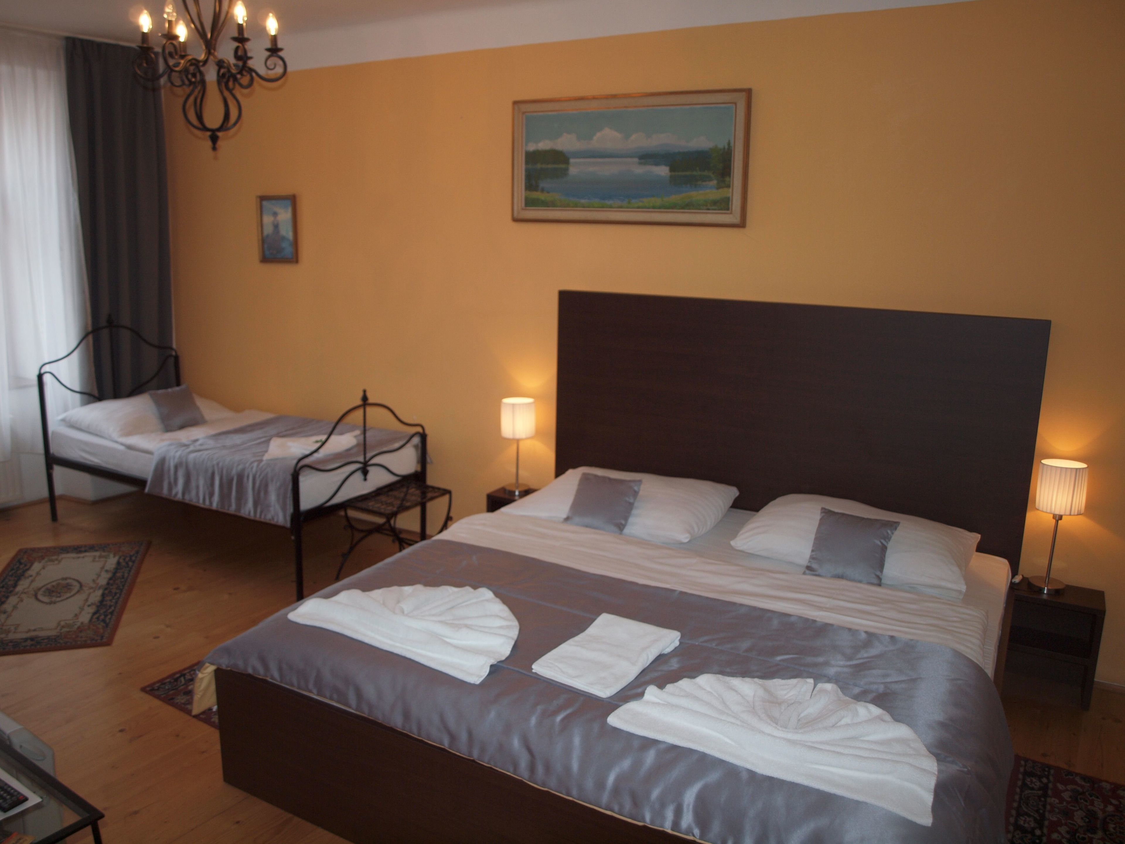 Une chambre d’hôtel avec un grand lit, deux petits lits, un lustre, un tableau et une table de chevet avec une lampe.