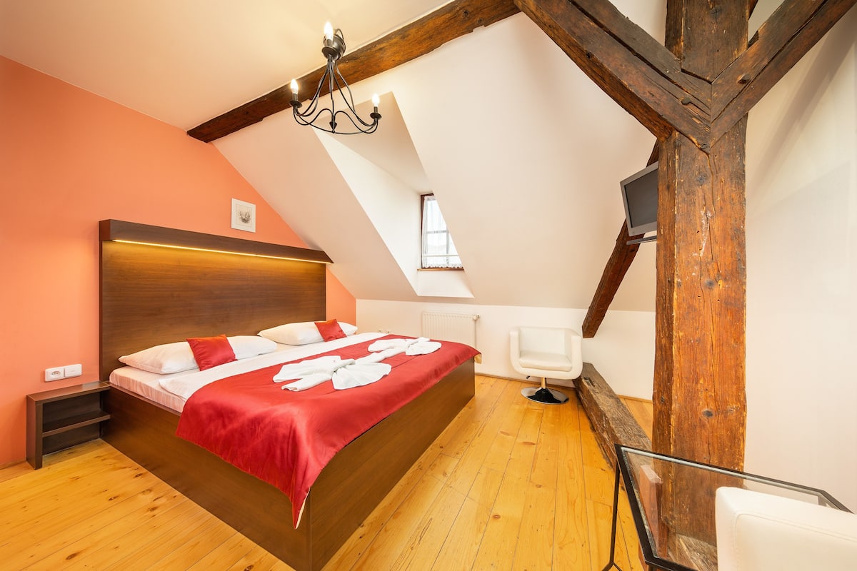 Un dormitorio con techo de madera, una cama con manta roja, una silla blanca y un televisor sobre una viga de madera.