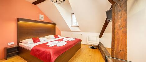 A bedroom with a wooden ceiling, a bed with a red blanket, a white chair, and a TV on a wooden beam.