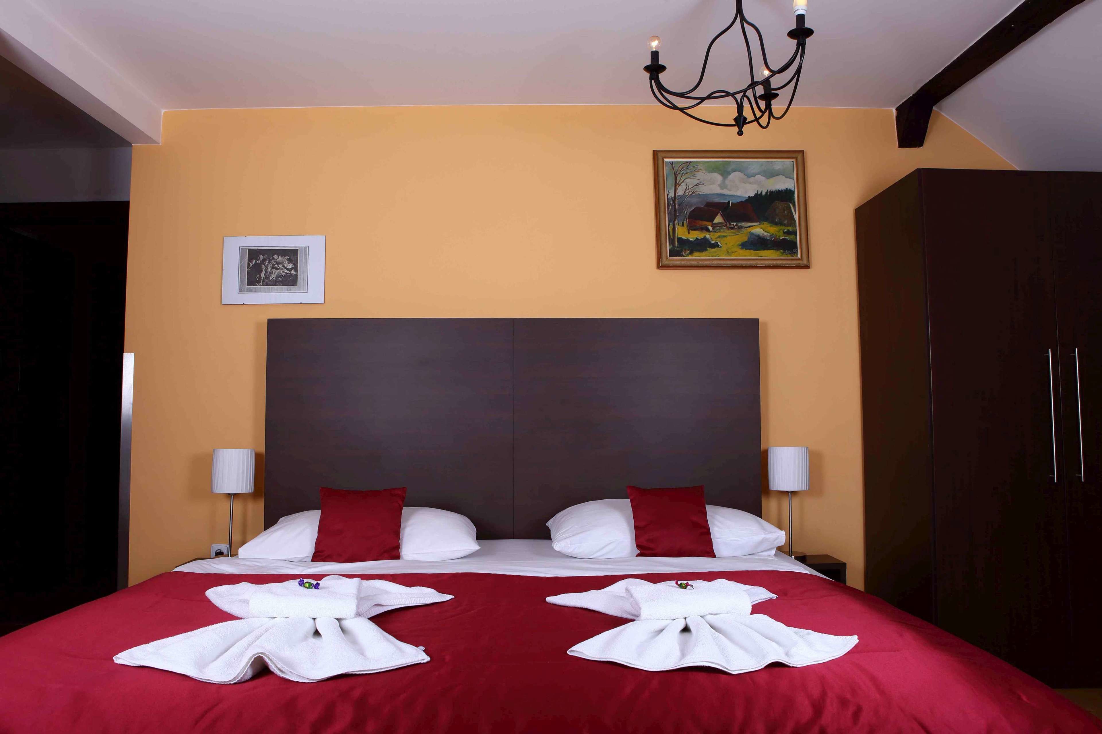 Habitación de hotel con una cama grande, dos almohadas rojas, una araña, un cuadro y un armario.