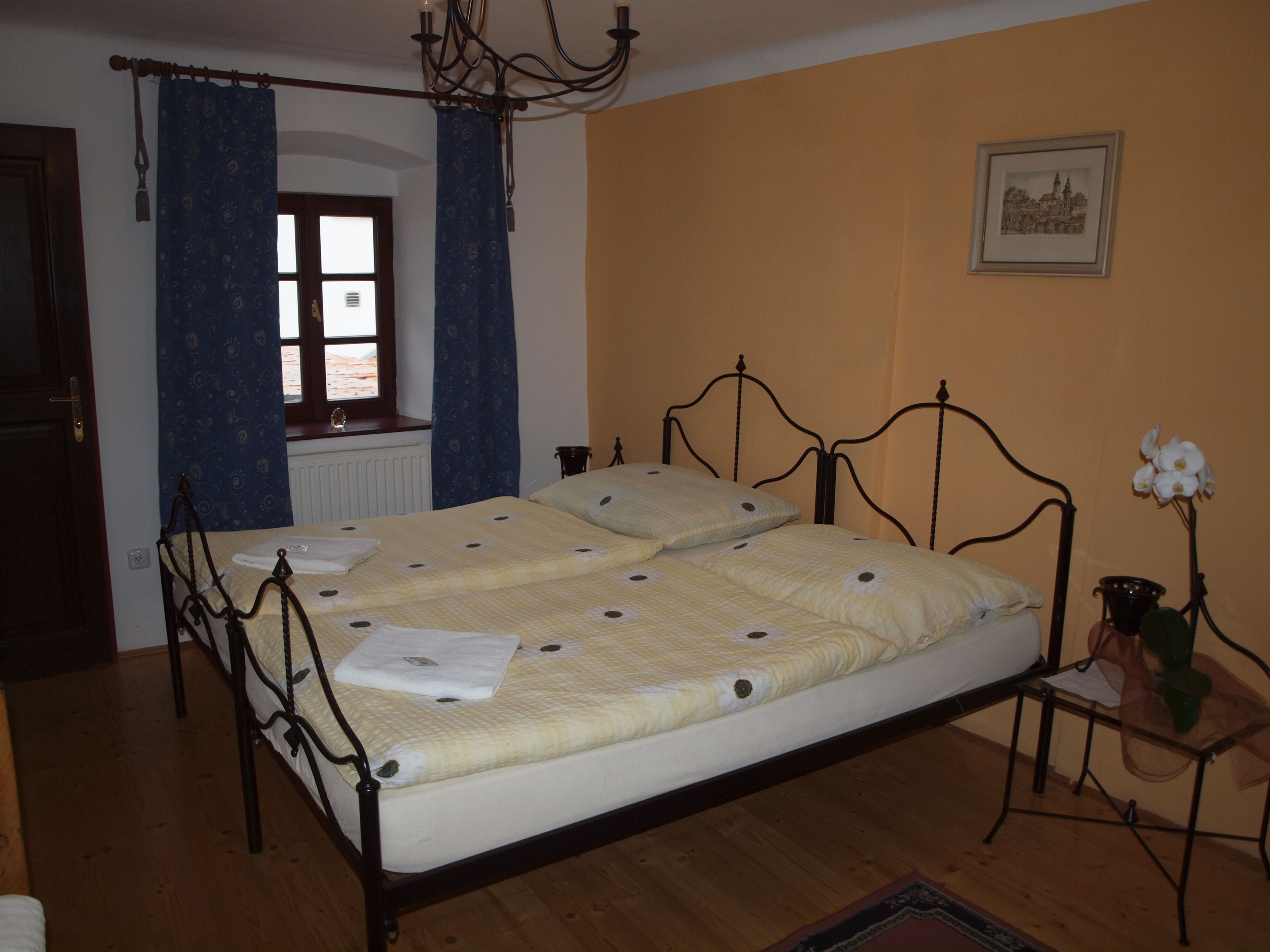 Une chambre à coucher avec un lit, une fenêtre avec des rideaux, un lustre, un tableau encadré au mur et une petite table sur laquelle se trouve un vase.