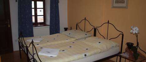 Un dormitorio con cama, ventana con cortinas, araña, cuadro en la pared y una mesita con un florero.