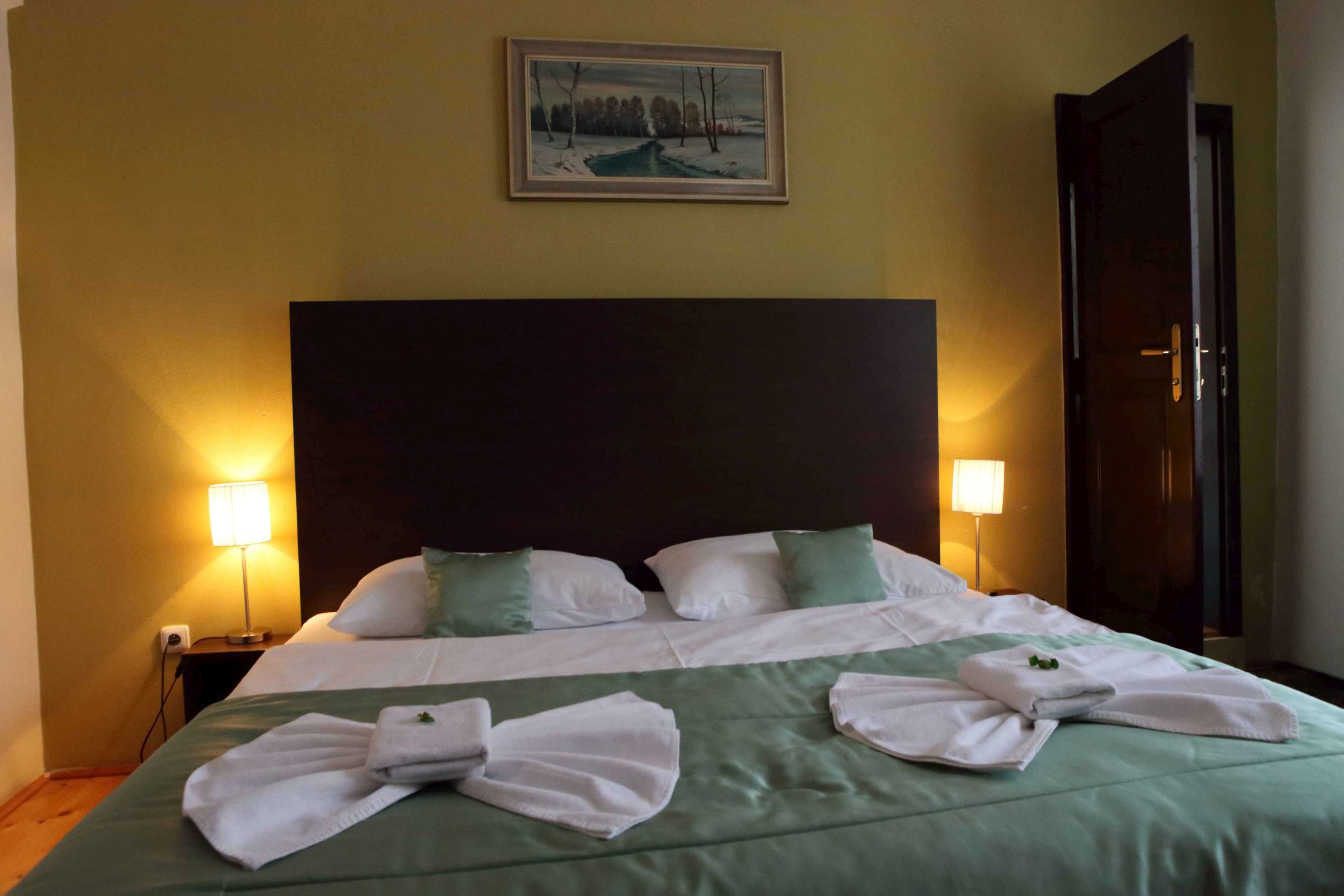 A hotel room with a large bed, two bedside lamps, a painting on the wall, and a door to the right.