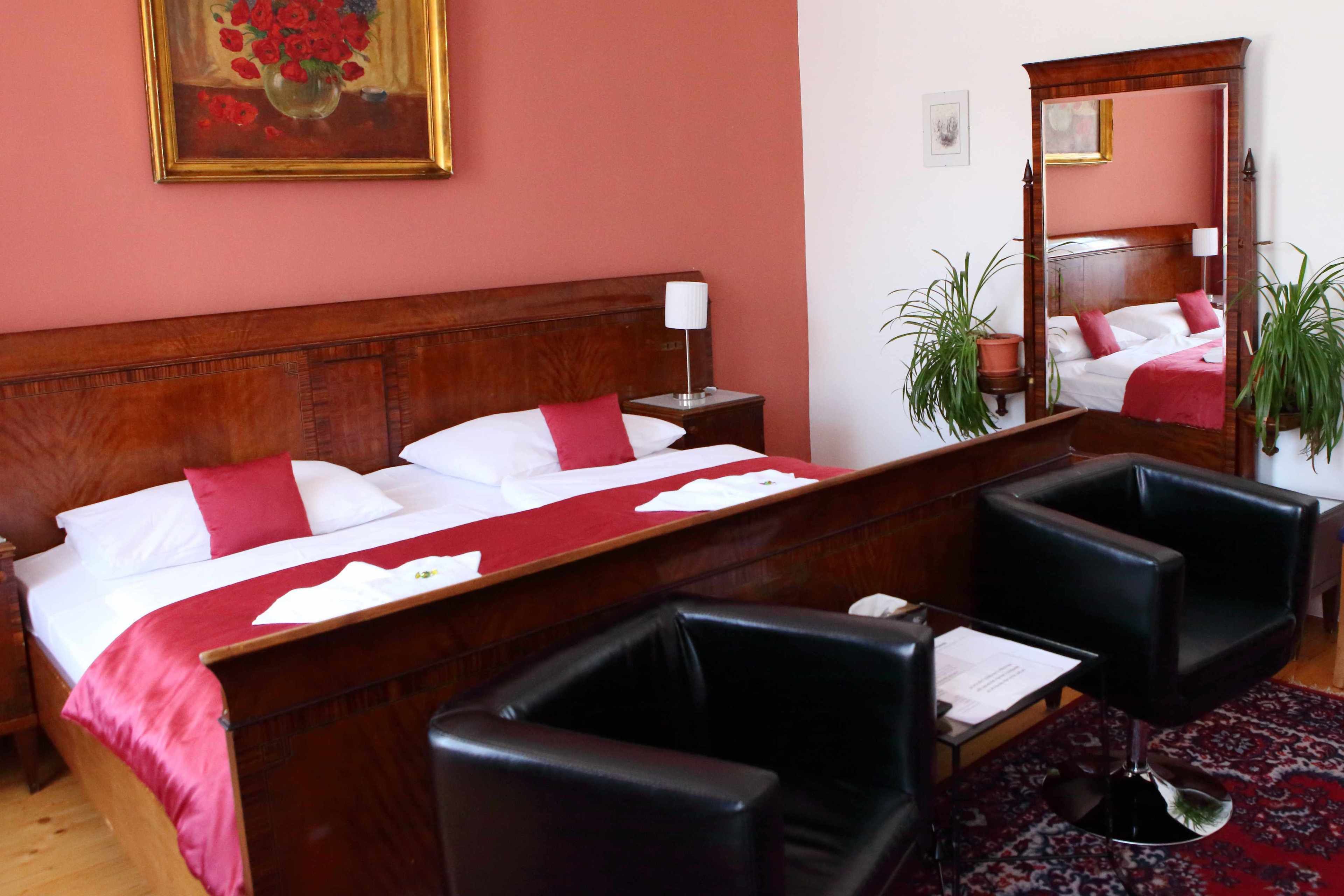 Quarto de hotel com uma cama de madeira, duas poltronas de couro preto, uma mesinha e um espelho.
