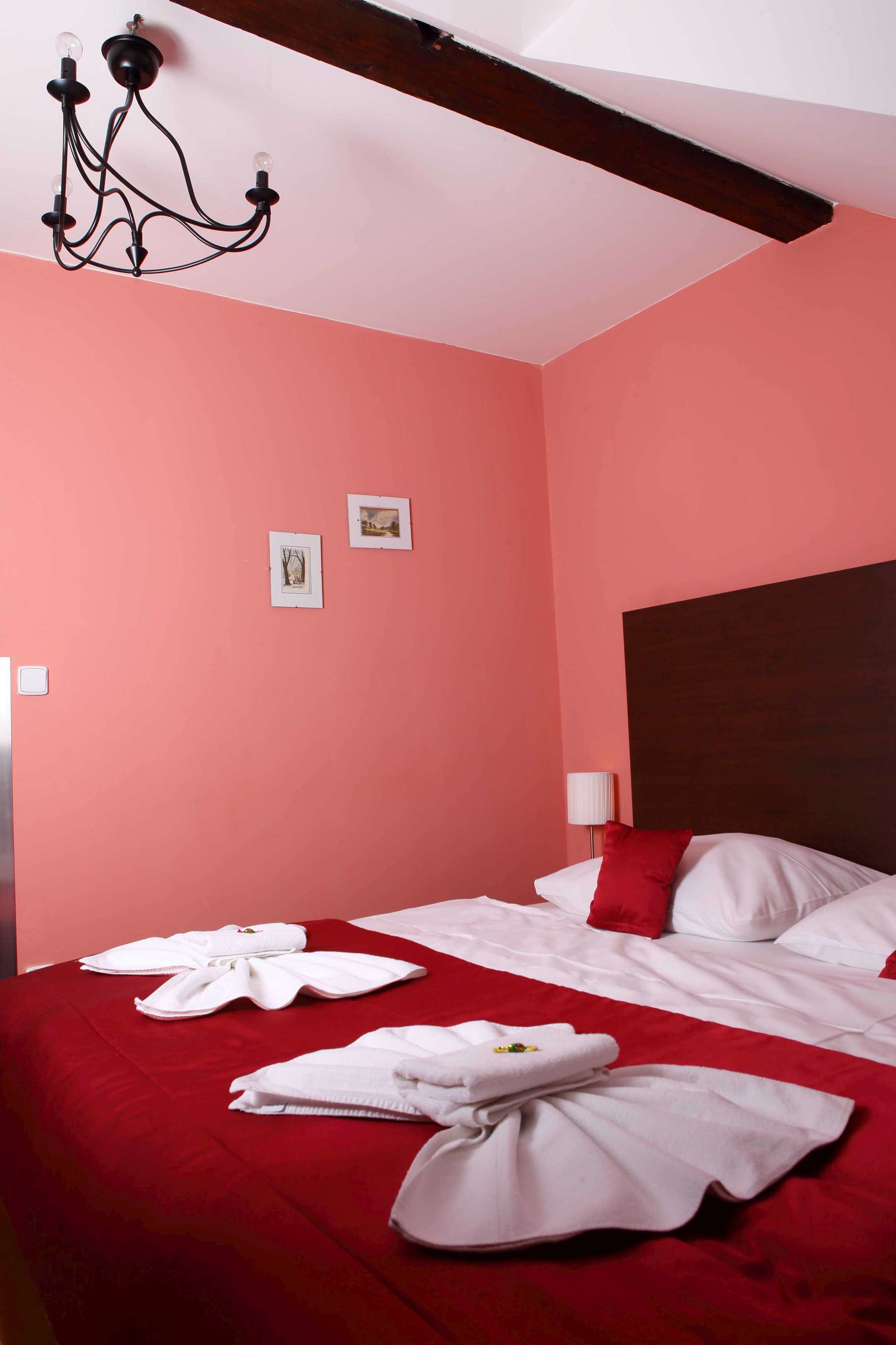 A hotel room with a red bedspread, white pillows, and a chandelier. There are two framed pictures on the wall and a bedside lamp.