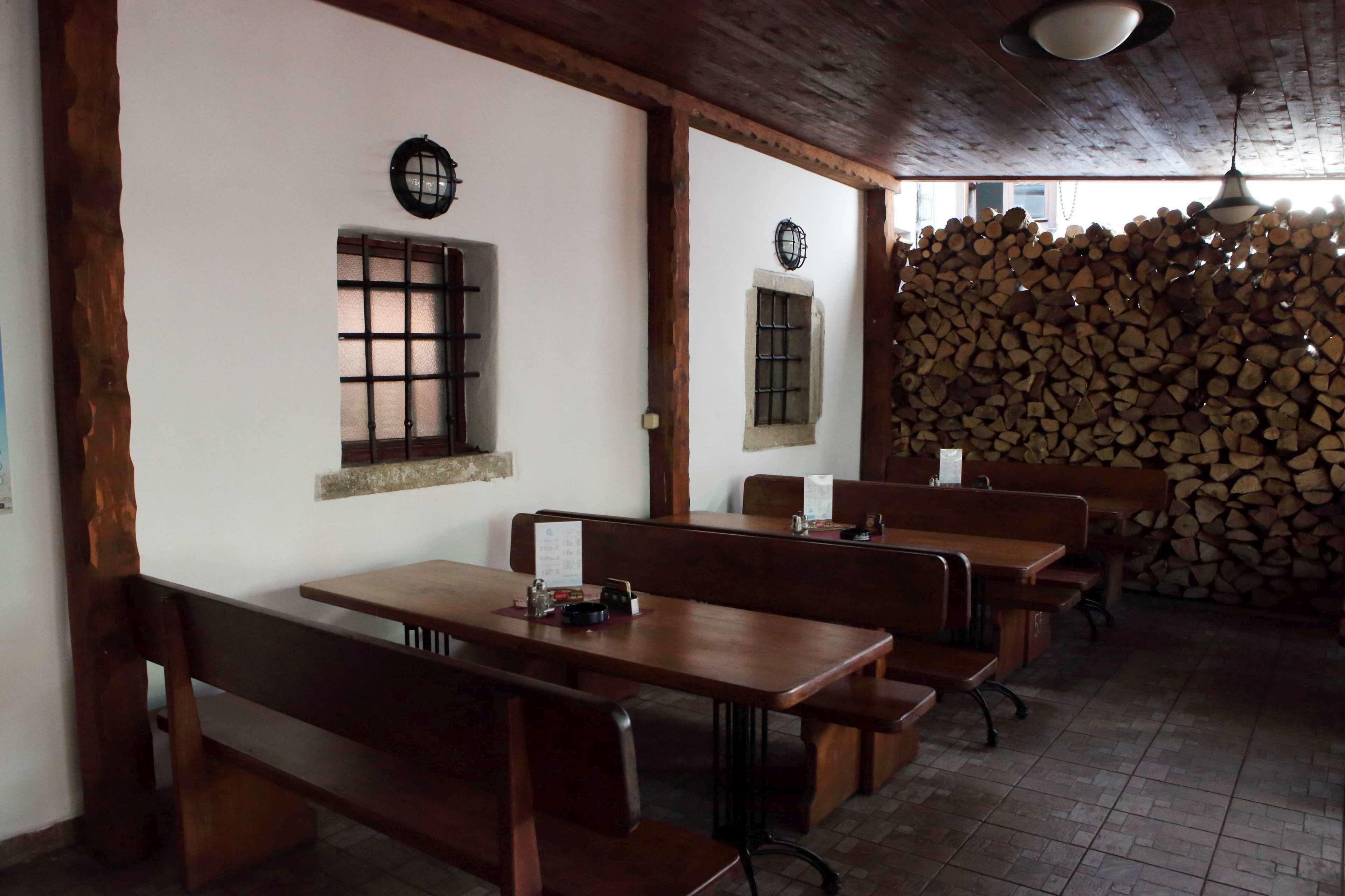 Interior rústic amb taules i bancs de fusta, un rellotge penjat a la paret i una pila de llenya.