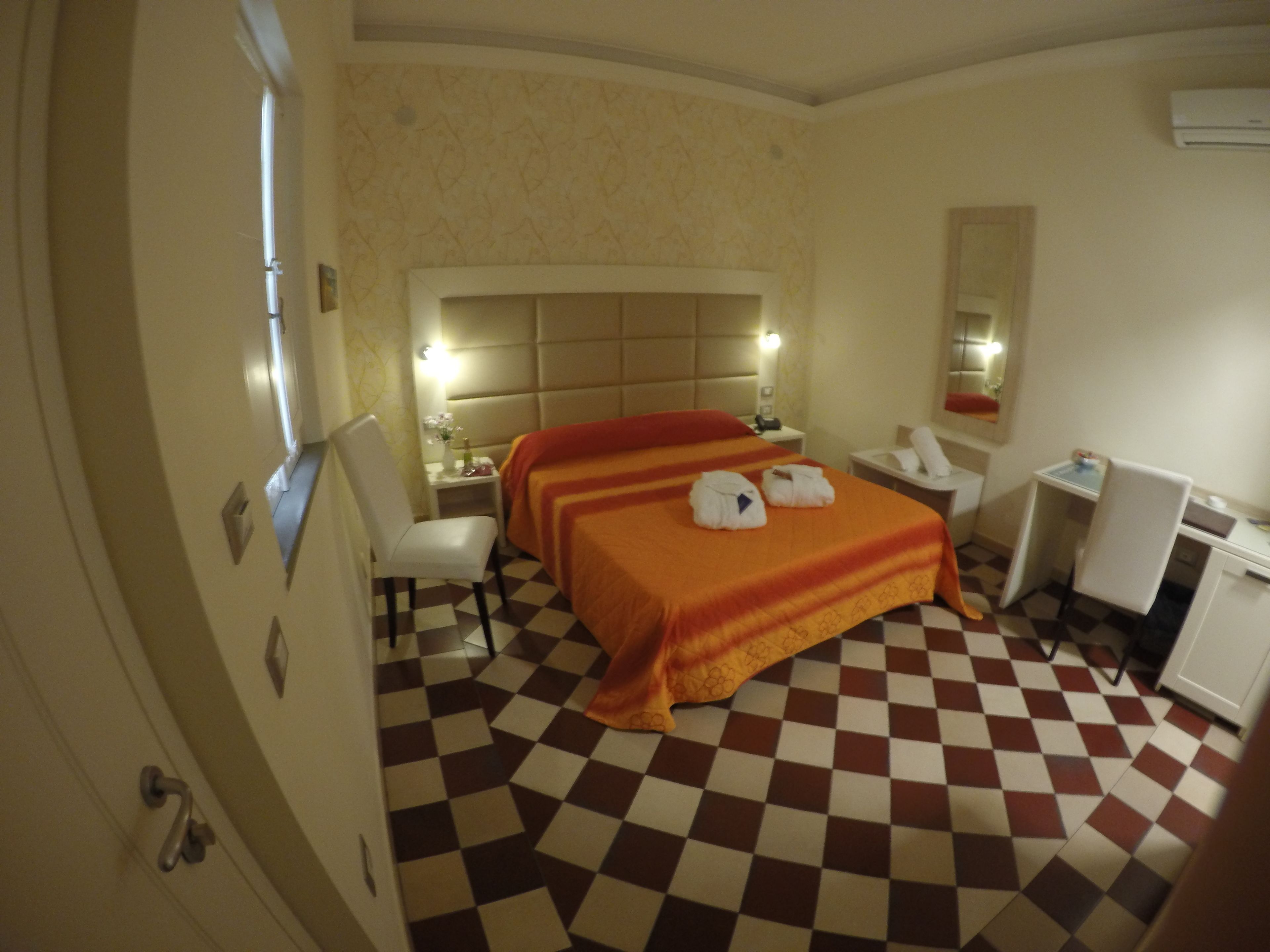 A hotel room with a checkered floor, a bed with an orange bedspread, a bedside table with a lamp, a chair, and a desk with a mirror.