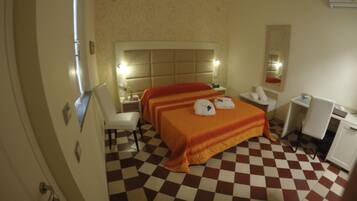 A hotel room with a checkered floor, a bed with an orange bedspread, a bedside table with a lamp, a chair, and a desk with a mirror.