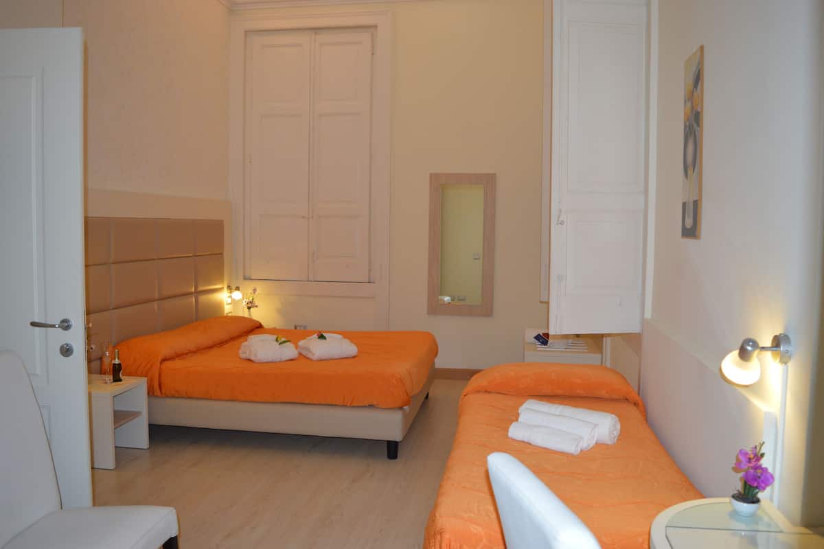 A hotel room with two beds, a bedside table with a lamp, a small table with a vase of flowers, and a door.