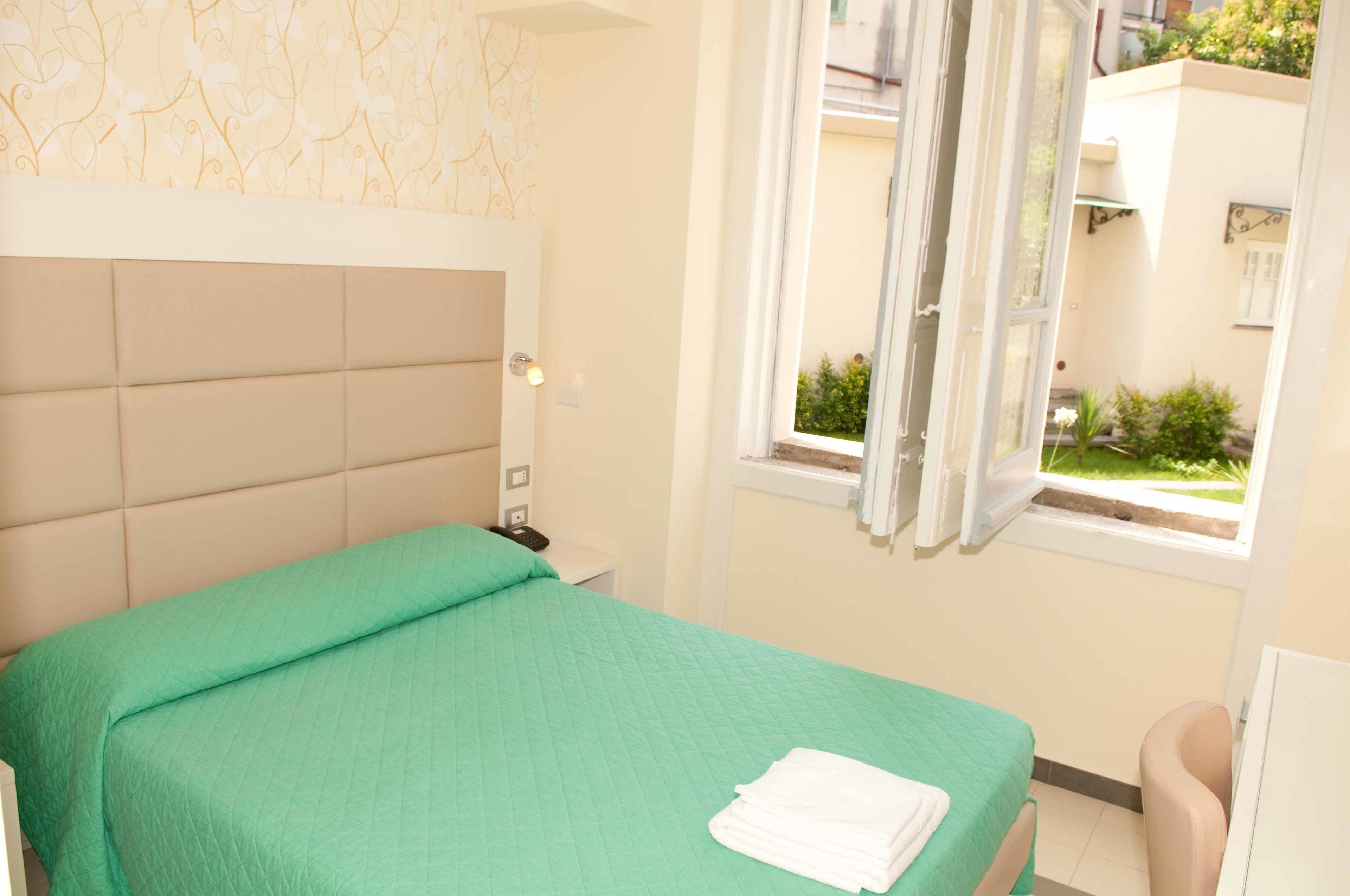 Quarto com colcha verde, cabeceira branca, mesa de cabeceira com telefone e janela com cortinas.