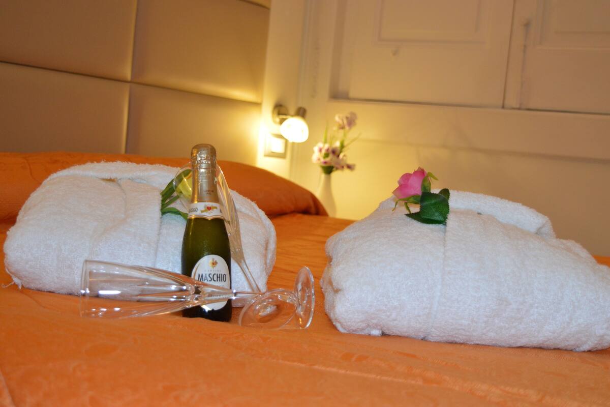 A bed with white towels, a bottle of Maschio wine, and a glass on a textured orange bedspread.