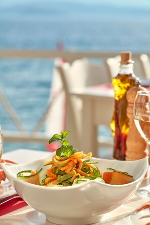 A white bowl of salad with a mint garnish, a bottle of olive oil, and a glass of wine on a table with a view of the sea. - Leda Village Resort (South Pelion)