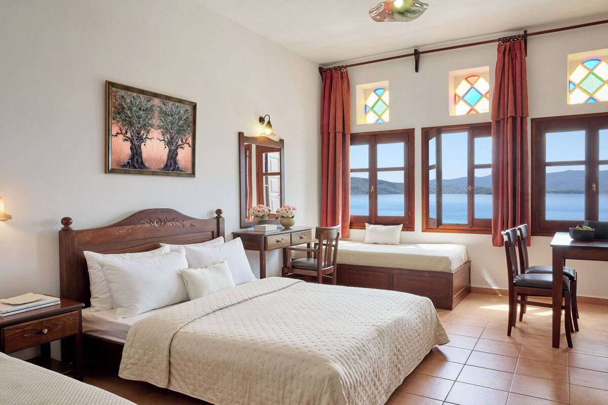 A hotel room with a large bed, a desk, and a chair. There are two windows with curtains, and a painting on the wall.